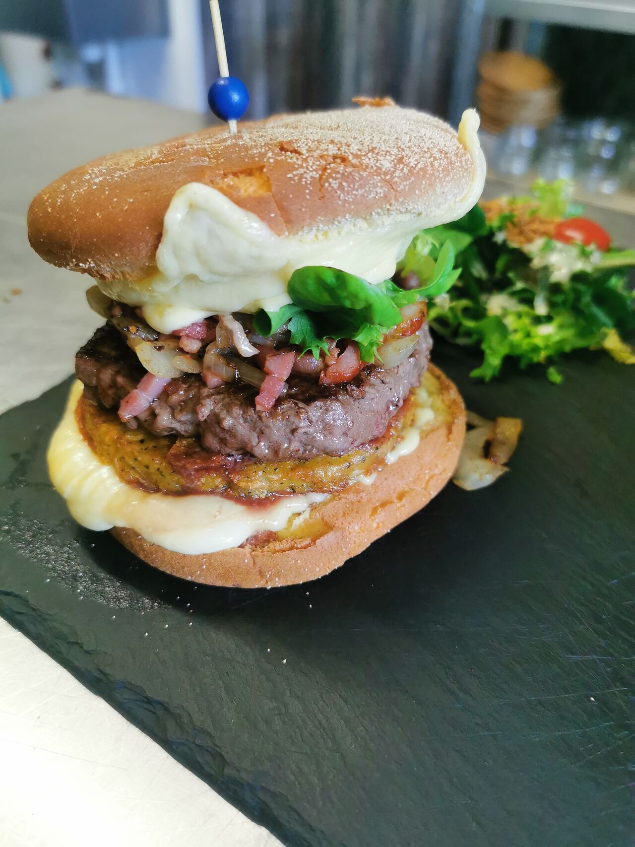 Le burger façon raclette. Galette de pomme de terre 150 g de viande de bœuf. Oignons et lardons caramélisés. Fromage à raclette
